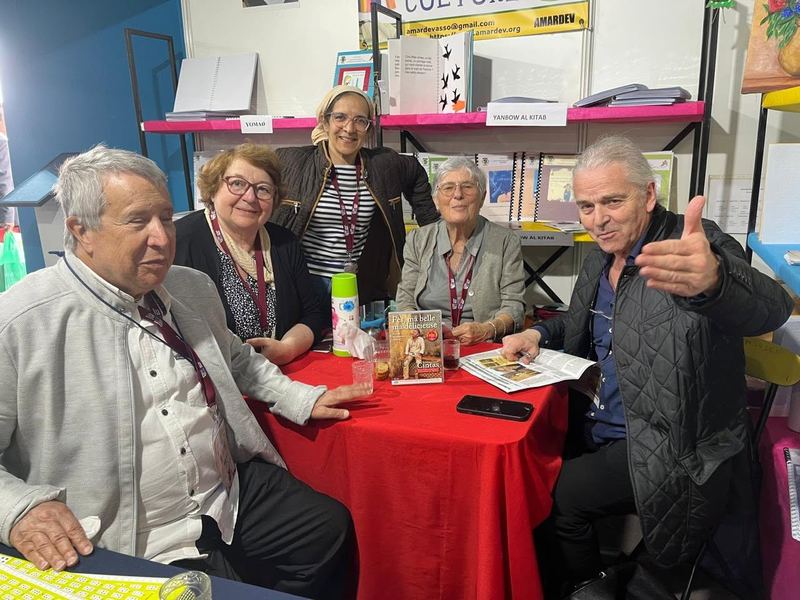 Un groupe de bénévoles et de visiteurs réunis autour d'une table au stand AMARDEV, avec un livre et des rafraîchissements