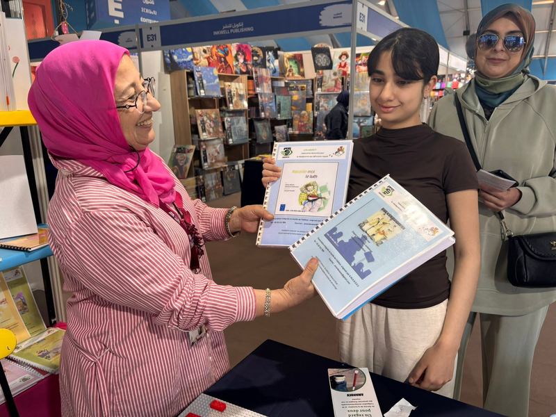 Une bénévole en foulard rose présente les livres « Raconte-moi le luth » et « Un regard pour deux » à une jeune visiteuse