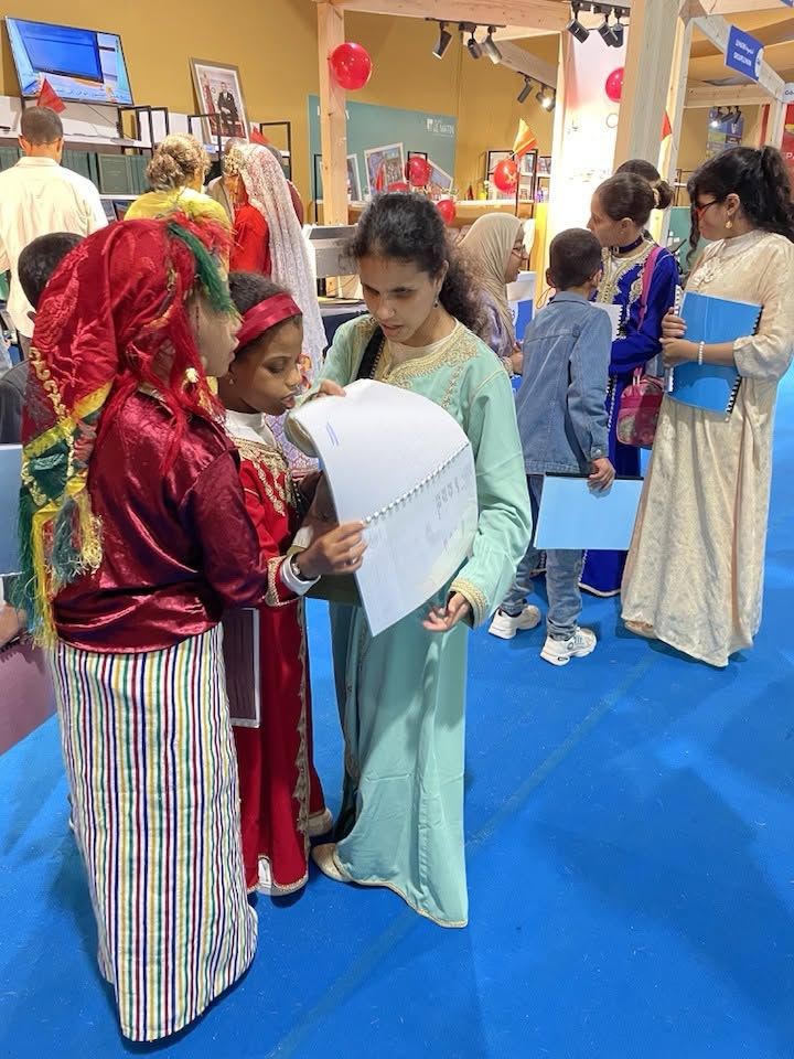 Trois jeunes filles en tenue traditionnelle examinent ensemble un livret au stand AMARDEV du SILEJ