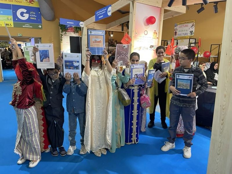 Un groupe d'enfants et de jeunes bénéficiaires d'AMARDEV, en costumes traditionnels marocains, lève fièrement des livrets reçus au stand du SILEJ