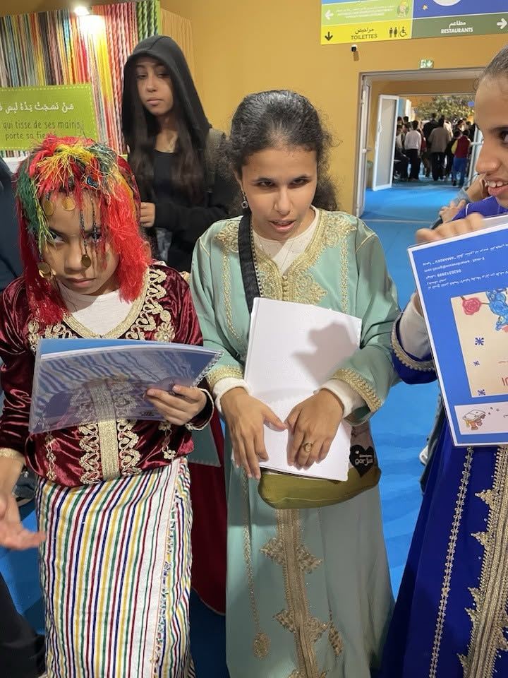 Deux jeunes filles en costume traditionnel consultent un livret, entourées d'autres visiteurs
